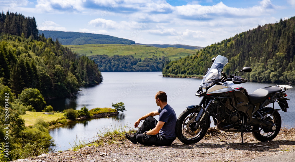 Adventure motorcycle and biker man traveling, sitting and watching landscape with lake and mountains, freedom travel lifestyle in Wales UK