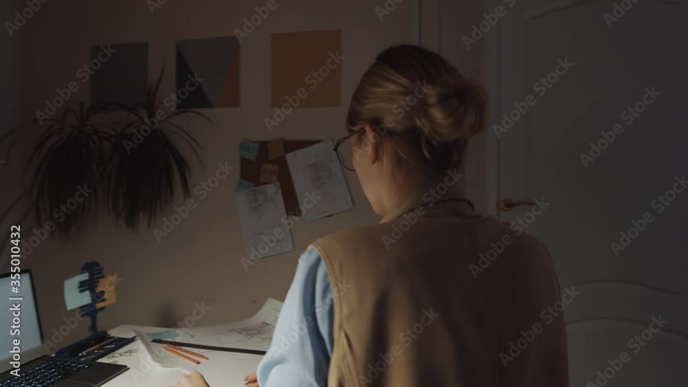 360-degree tracking shot of young female architect using laptop at desk, then walking to shelves, taking file and checking construction plans while working late in dark office 360-degree tracking shot of young female architect using laptop at desk, then walking to shelves, taking file and checking construction plans while working late in dark office