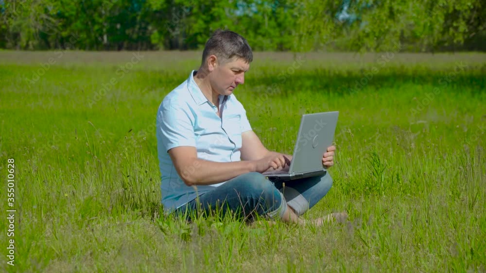 Man Works behind a Laptop Sitting on the Grass At The Sunny Day. Freelancer Remote Work Concept. 4K Man Works behind a Laptop Sitting on the Grass At The Sunny Day. Freelancer Remote Work Concept. 4K