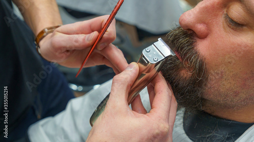 Barber trimming bearded man with shaving machine in barbershop. Hairstyling process. Haircut concept.