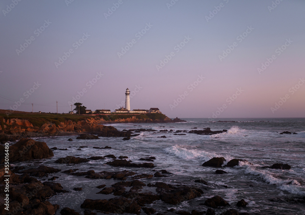 White lighthouse on the mountain coast of the ocean at sunset. Rocky beach with waves in the evening of summer against the background of a purple evening sky without clouds