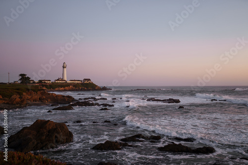 Landscape of a white lighthouse with buildings against the backdrop of purple sunset on the Pacific rocky coast with powerful waves, green grass, stones and a beautiful sky with clouds.