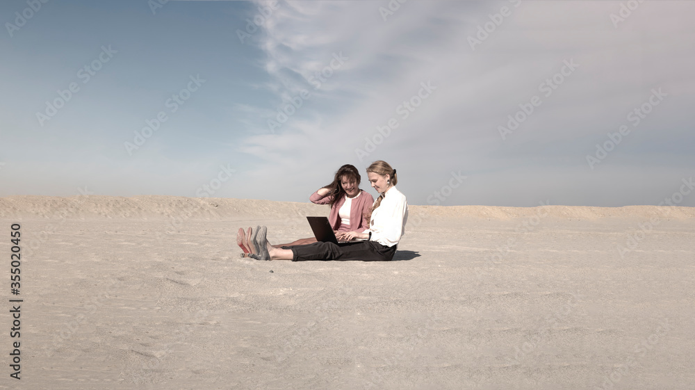 two women in office clothes sit on the sand in the desert with smiling faces looking at laptop