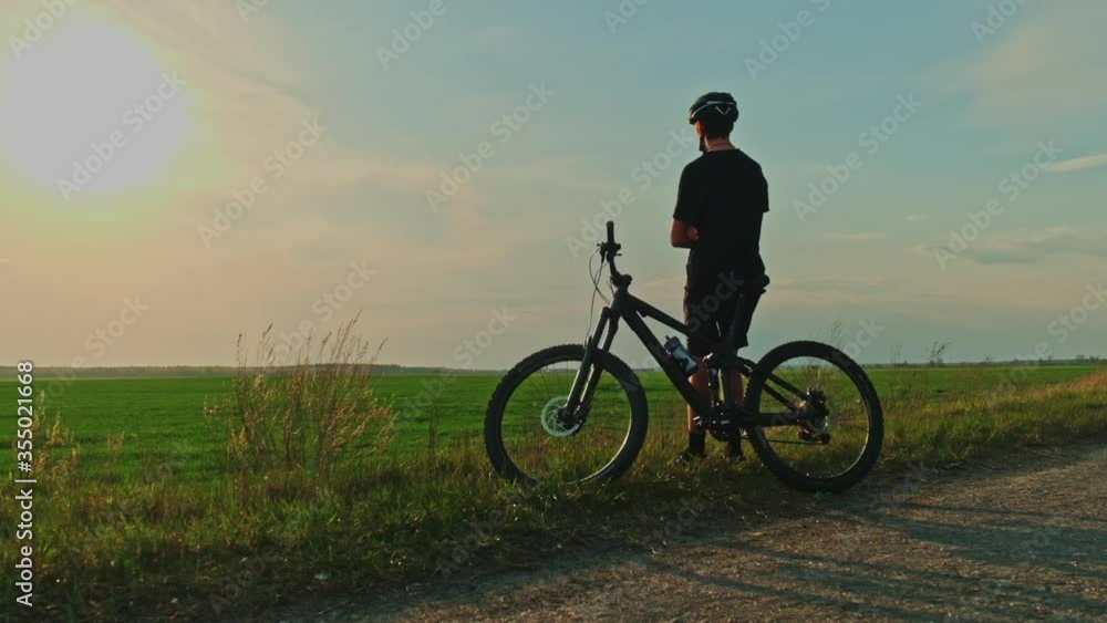 Cyclist enjoys the sunset. A young man leaning on a Bicycle looks at the evening sunset against the background of a green farm field. Young cyclist looking into the distance during sunrise Cyclist enjoys the sunset. A young man leaning on a Bicycle looks at the evening sunset against the background of a green farm field. Young cyclist looking into the distance during sunrise