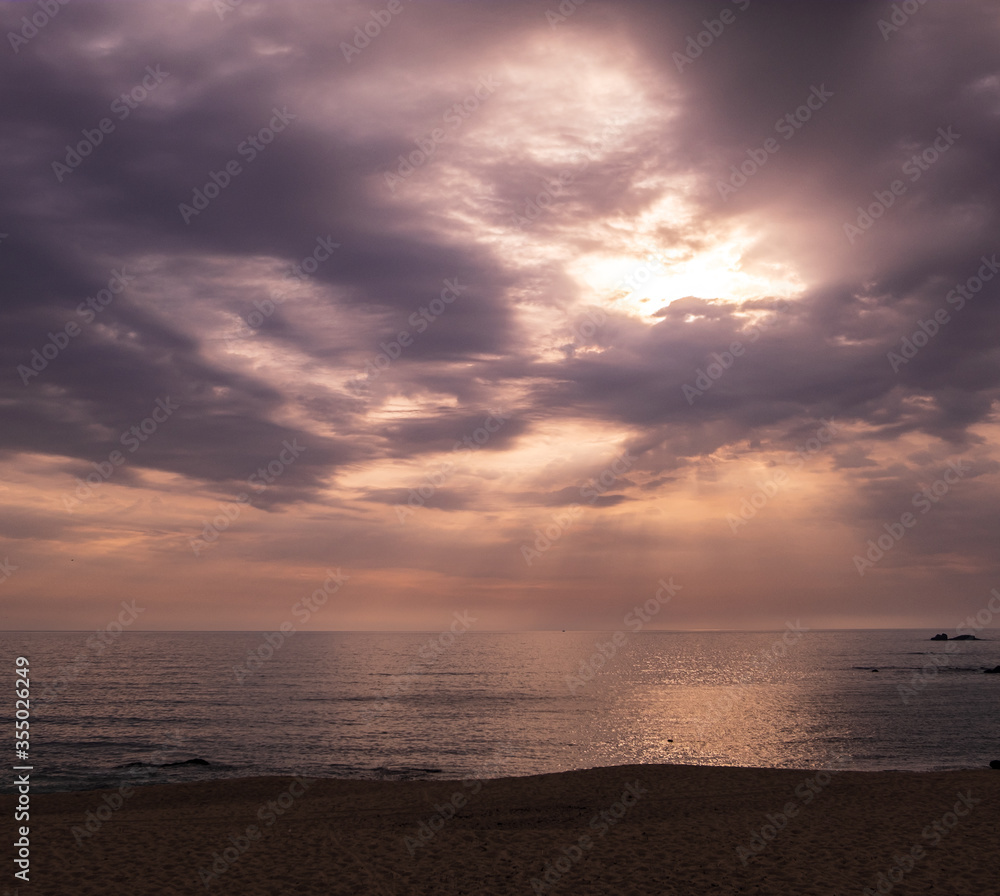 Beach at sunset with rays of sunlight shining through clouds onto ocean.