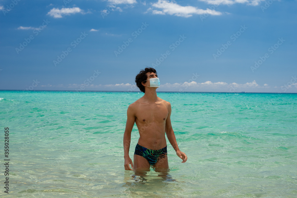 Young handsome man in paradisiac beach coming out of the ocean with surgery protective mask on sunny day during 2020 coronavirus summer holiday Young handsome man in paradisiac beach coming out of the ocean with surgery protective mask on sunny day during 2020 coronavirus summer holiday