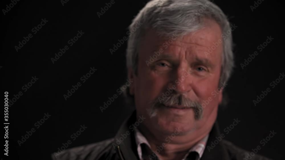 Handsome grey haired senior man looking at camera and smile with camera slide out wearing jacket in semi lit light studio isolated on black background. Prores 422. Handsome grey haired senior man looking at camera and smile with camera slide out wearing jacket in semi lit light studio isolated on black background. Prores 422.