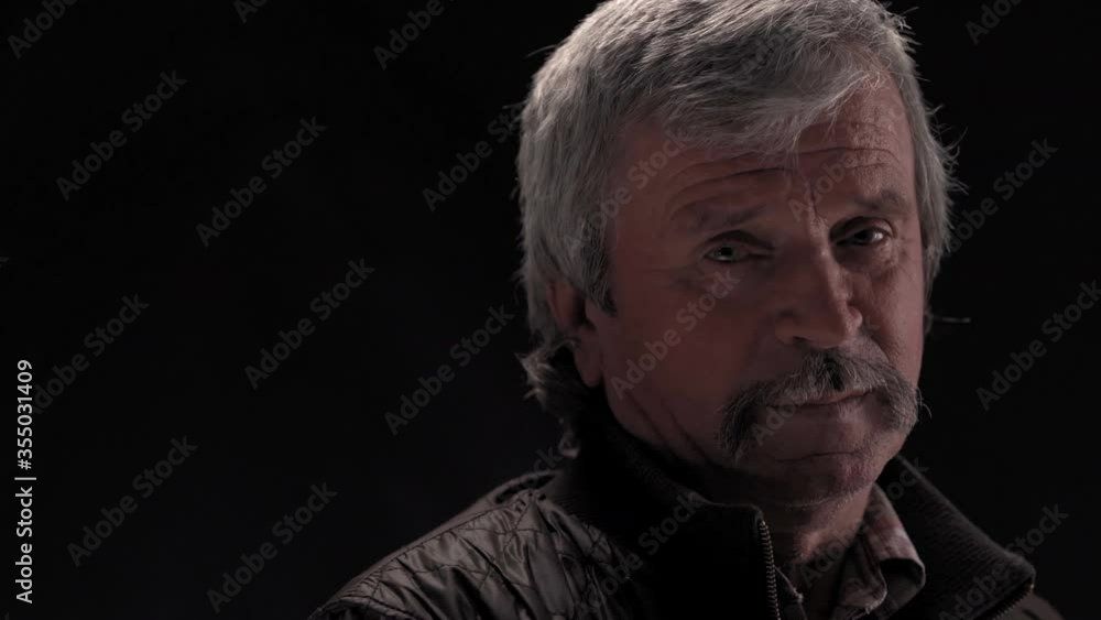 Handsome grey haired senior man looking at camera wearing jacket in semi lit light studio isolated on black background. Prores 422. Handsome grey haired senior man looking at camera wearing jacket in semi lit light studio isolated on black background. Prores 422.