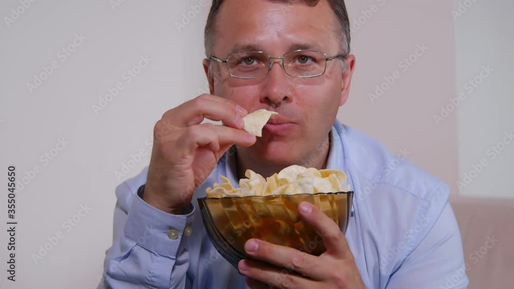 Caucasian young man best sitting on couch at home, eating potato chips. Guy watching sport channel on TV with football game. Sport fan time spending concept. Caucasian young man best sitting on couch at home, eating potato chips. Guy watching sport channel on TV with football game. Sport fan time spending concept.