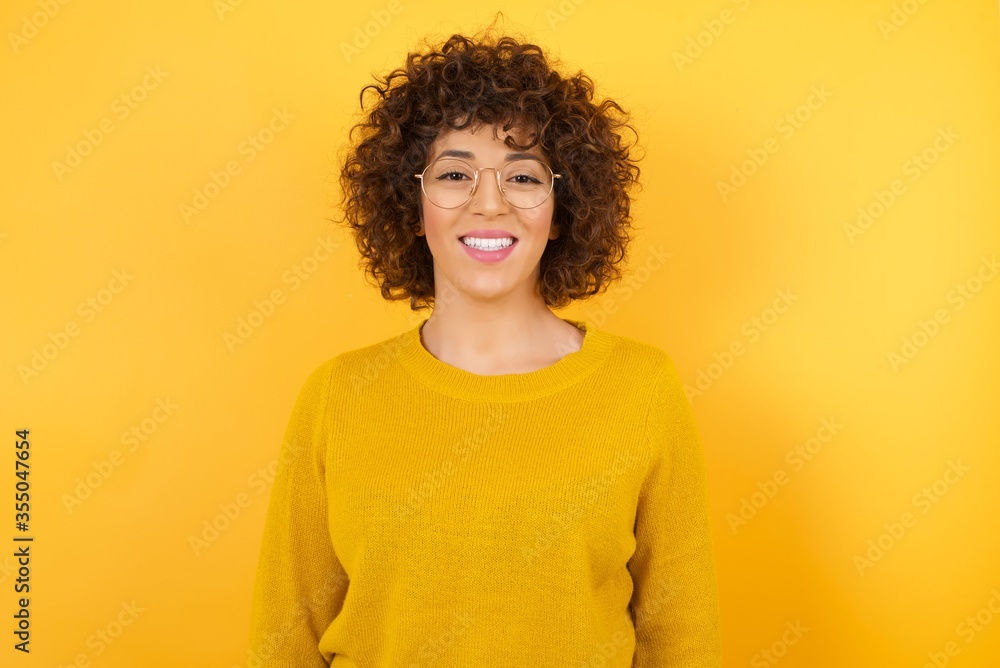 Young beautiful business woman with curly hair wearing yellow sweater over isolated yellow background with a happy and cool smile on face. Lucky person. Young beautiful business woman with curly hair wearing yellow sweater over isolated yellow background with a happy and cool smile on face. Lucky person.