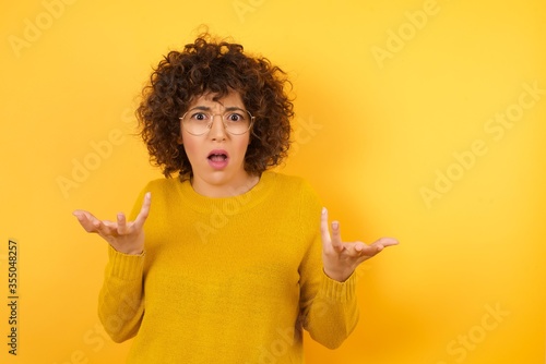 Frustrated Arab female feels puzzled and hesitant, shrugs shoulders in bewilderment, keeps mouth widely opened, doesn't know what to do, isolated over yellow background. Studio shot.