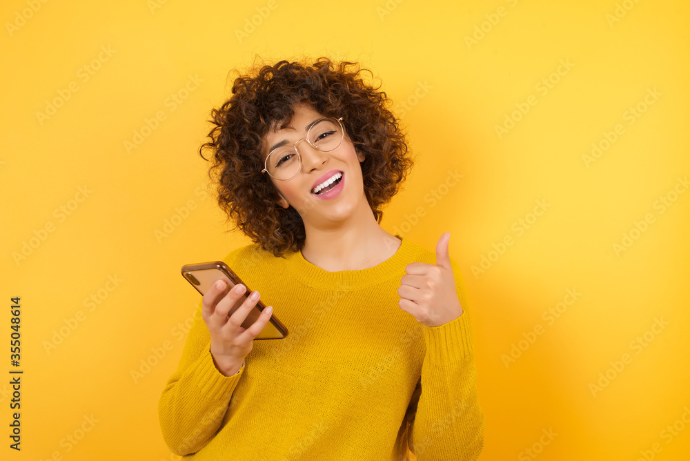 Portrait of beautiful Arab businesswoman wearing yellow sweater using and texting with smartphone over isolated background happy with big smile doing ok sign, thumb up with fingers, excellent sign Portrait of beautiful Arab businesswoman wearing yellow sweater using and texting with smartphone over isolated background happy with big smile doing ok sign, thumb up with fingers, excellent sign