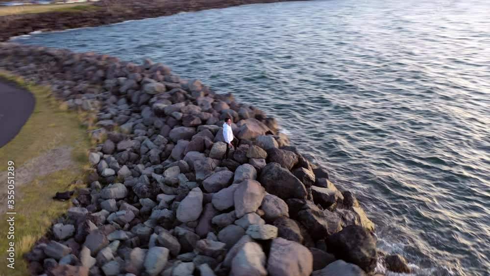 Aerial panning shot of man standing on rocks by sea during sunset, drone flying around tourist on groyne - Reykjavík, Iceland Aerial panning shot of man standing on rocks by sea during sunset, drone flying around tourist on groyne - Reykjavík, Iceland