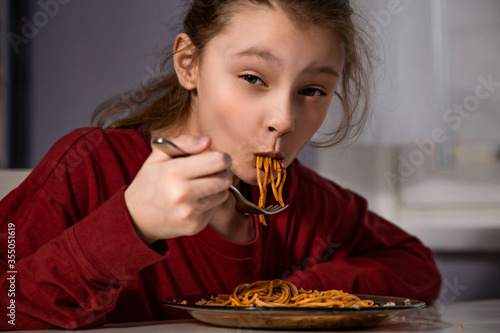 Pretty enjoying grimacing kid girl eating tasty spaghetti on the dinner on the home kitchen. Closeup