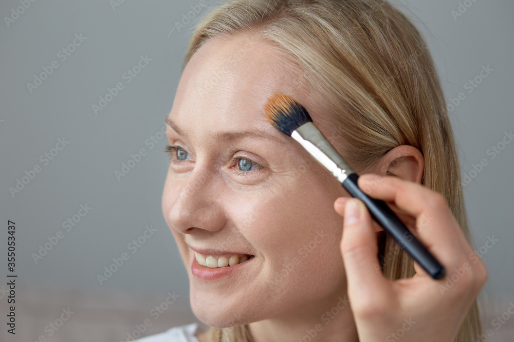A smiling young girl puts a tonal cream on her face with a brush.