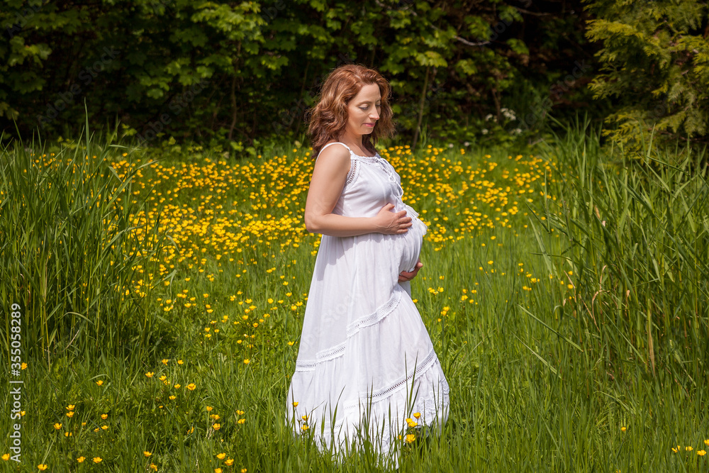 Beautiful pregnant woman in white dress standing in the green park in sunny day. Beautiful pregnant woman in white dress standing in the green park in sunny day.