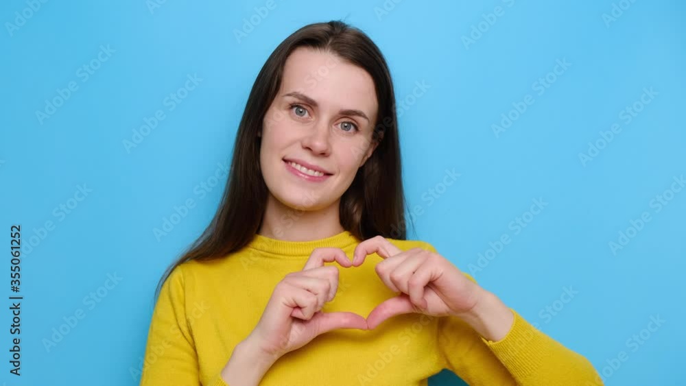 Portrait of glad beautiful young woman shows heart gesture over chest, being passionate, express love to close person, wears yellow jumper, stands over blue studio background. Sincere confession Portrait of glad beautiful young woman shows heart gesture over chest, being passionate, express love to close person, wears yellow jumper, stands over blue studio background. Sincere confession