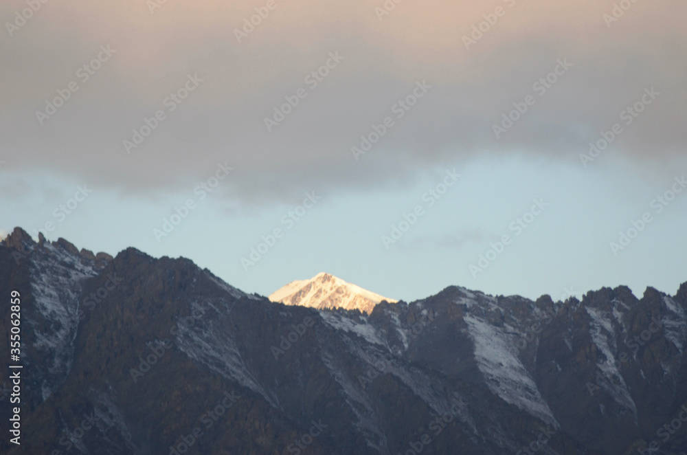 Sunlight is falling on a snowy mountain peak in the Western Himalayas. Rocky mountain slopes are in the foreground. Heavy clouds are touched with the colours of sunrise.