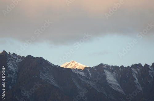Sunlight is falling on a snowy mountain peak in the Western Himalayas. Rocky mountain slopes are in the foreground. Heavy clouds are touched with the colours of sunrise. © Anne