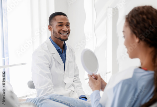 Black lady checking beautiful smile in mirror, dentist watching her