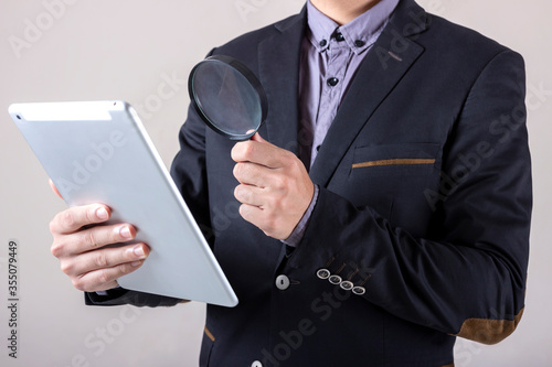 Man in business suit holding a magnifying glass looking through a tablet, concept of researching, examining and analyzing.