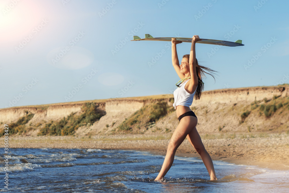 Obraz premium Young caucasian woman surfer in white shirt going to the sea with her surfboard above her head.