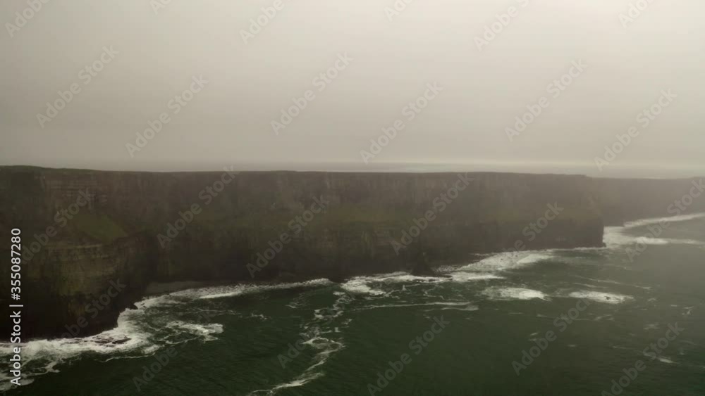 Aerial panning shot of rocky cliff in sea during foggy weather, drone flying over waves splashing in ocean against sky - Southwestern Iceland, Ireland Aerial panning shot of rocky cliff in sea during foggy weather, drone flying over waves splashing in ocean against sky - Southwestern Iceland, Ireland