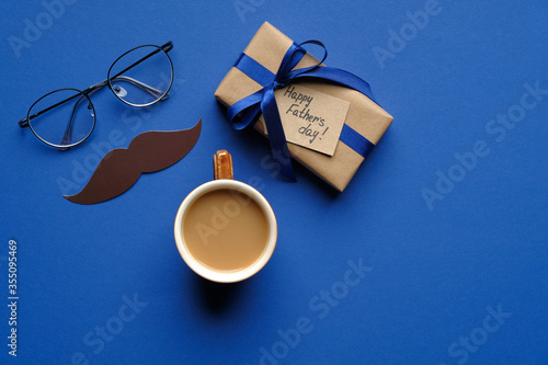 Happy Father's day concept. Top view gift box wrapped blue ribbon, greeting card, cup of coffee, mustache and glasses on blue table. Fathers day banner mockup.