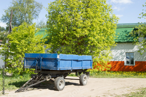 A blue empty trailer is located in a sunny meadow near a metal modern bright orange barn or agricultural building, warehouse. Green trees and bushes.
