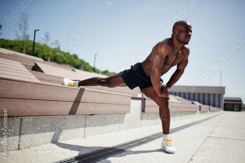 Darkskinned athlete with a naked torso does an exercise to stretch the internal muscles of the thighs leaning on a bench outdoor.