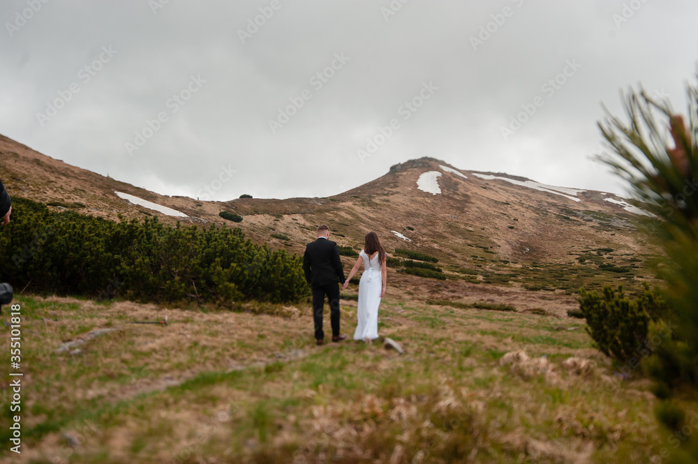 Back view shot of young wedding couple of groom and bride in white dress holding hands while walking on the top of the hill in mountains 