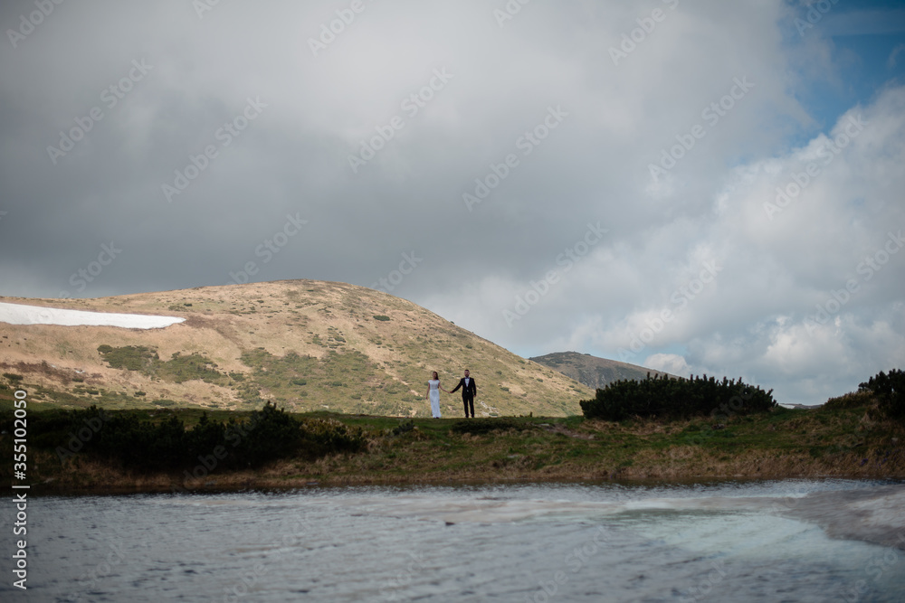 Scenic view of beautiful spring mountains and a lake. Wedding couple holding hands and walking on a hill Scenic view of beautiful spring mountains and a lake. Wedding couple holding hands and walking on a hill