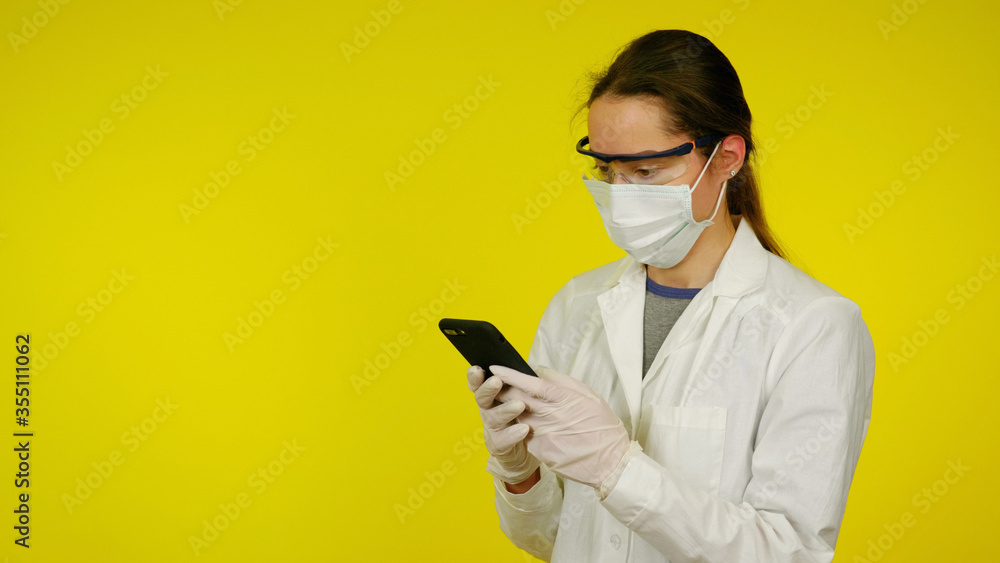 Young doctor in a protective medical mask, glasses and latex gloves looks at the smartphone. Nurse in a white coat on yellow background is looking for information in a smartphone