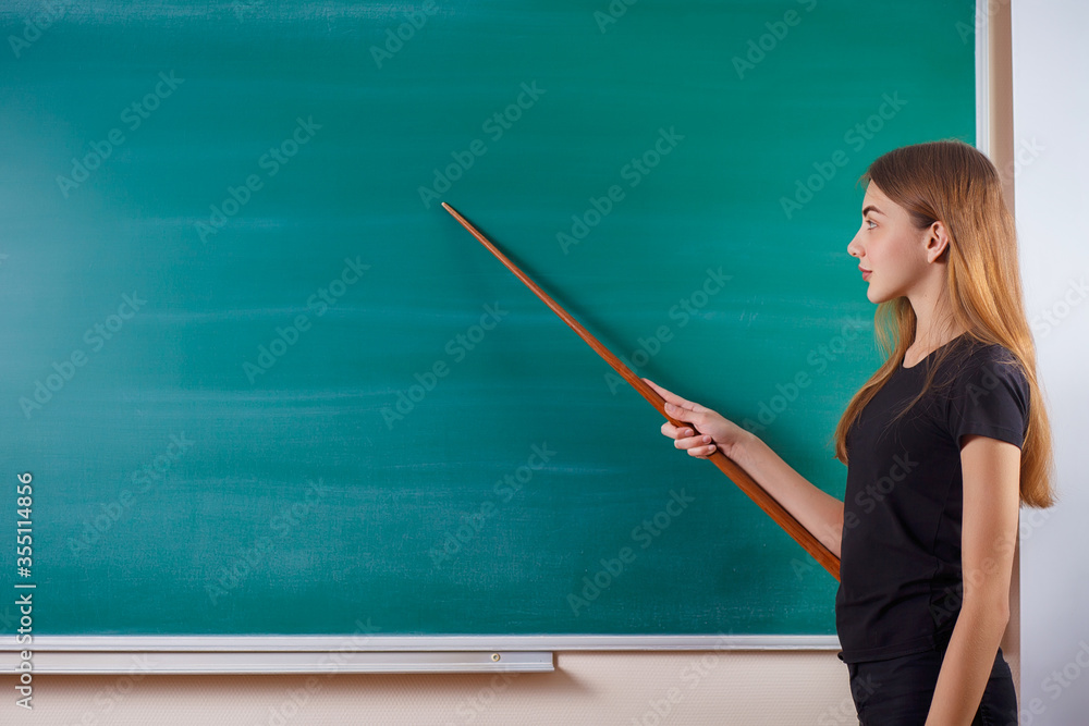 Young attractive student at the school board. She rejoices at the beginning of the school year and holds a pointer in her hands. Back to school. Copy space Young attractive student at the school board. She rejoices at the beginning of the school year and holds a pointer in her hands. Back to school. Copy space