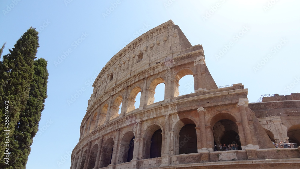Rome Italy Colosseum July 2017 midday in fine weather with blue sky, large cypress trees next to the building