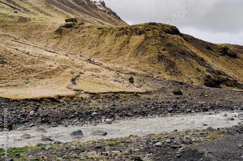 A shallow mountain river flows against a mountain with yellow grass in Iceland.