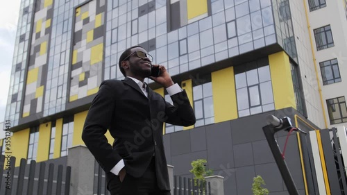 Handsome black businessman in a black suit talking on the phone outdoors on the background of a glass office center, next to him is an electric scooter.