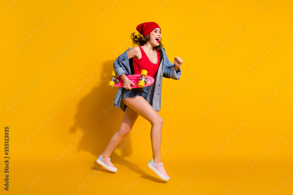 Full size profile side photo energetic beautiful girl student jump hold longboard excited run skate board sporty workout wear red headwear jeans tank-top isolated bright color background