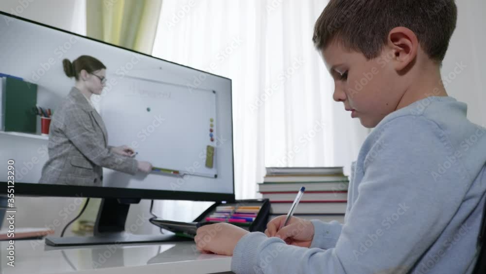 electronic training, smart elementary school student learns lessons at home sitting at computer and watches video broadcast of lesson with teacher on monitor screen electronic training, smart elementary school student learns lessons at home sitting at computer and watches video broadcast of lesson with teacher on monitor screen
