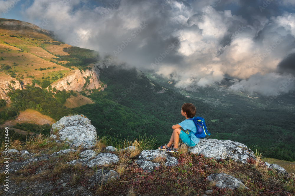 Cute little boy with blue backpack sits on a cliff above a huge mountain valley and looks at the mountains with beautiful powerful clouds. Trekking with kids, family travelling. Childhood in nature.