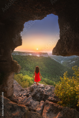Beautiful young girl with long hair in a red dress looks at the sunset, standing on the edge of a cliff with a fancy arch. Stunning sunset view from an artificial cave.