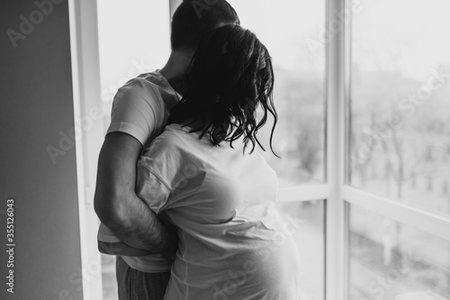 Pregnant couple cuddling each other at home. Man holding pregnant woman's stomach in bedroom. Pregnant young woman are waiting for a child at home by the window. Loving family, husband and wife.