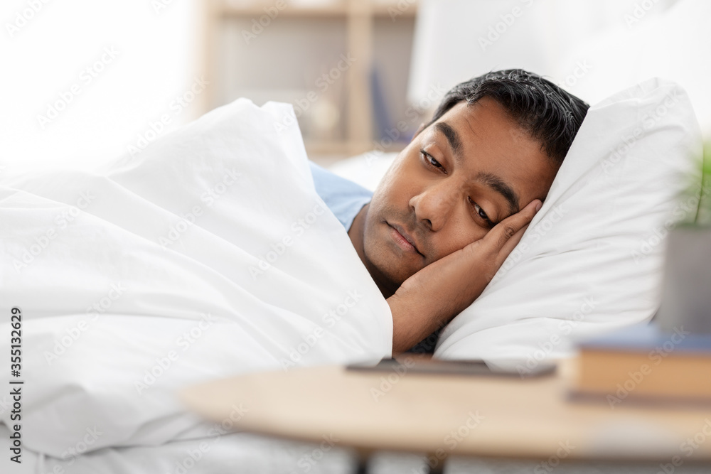 people, bedtime and rest concept - sleepy indian man in bed looking at smartphone lying on table at home people, bedtime and rest concept - sleepy indian man in bed looking at smartphone lying on table at home