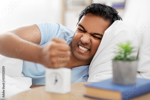 people, bedtime and rest concept - angry indian man with alarm clock lying in bed at home