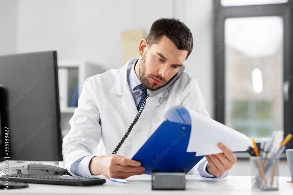 healthcare, medicine and people concept - male doctor with clipboard calling on desk phone at hospital