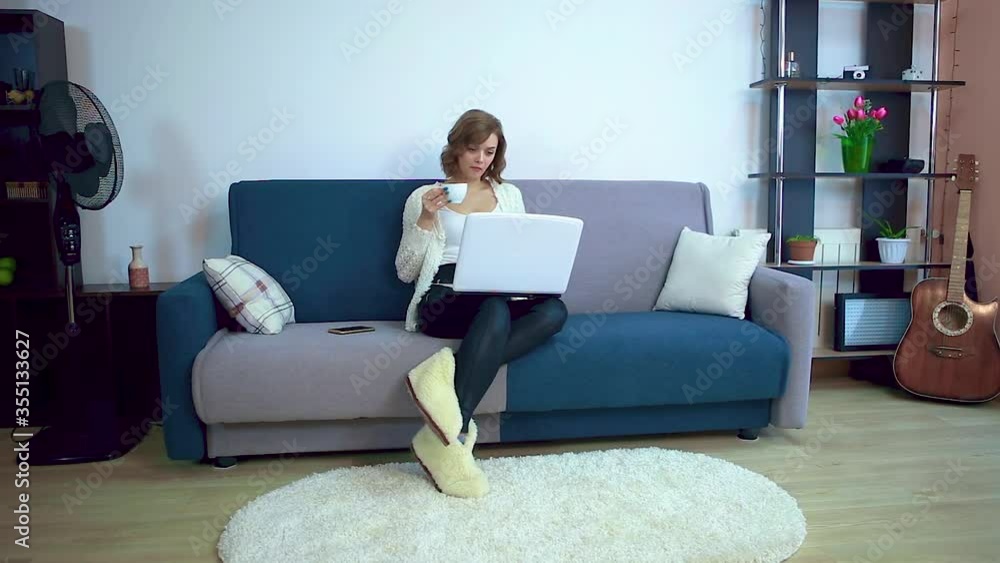 A girl in a white sweater is drinking hot tea or coffee from a white cup while sitting on a sofa, working on a laptop. A girl in a white sweater is drinking hot tea or coffee from a white cup while sitting on a sofa, working on a laptop.