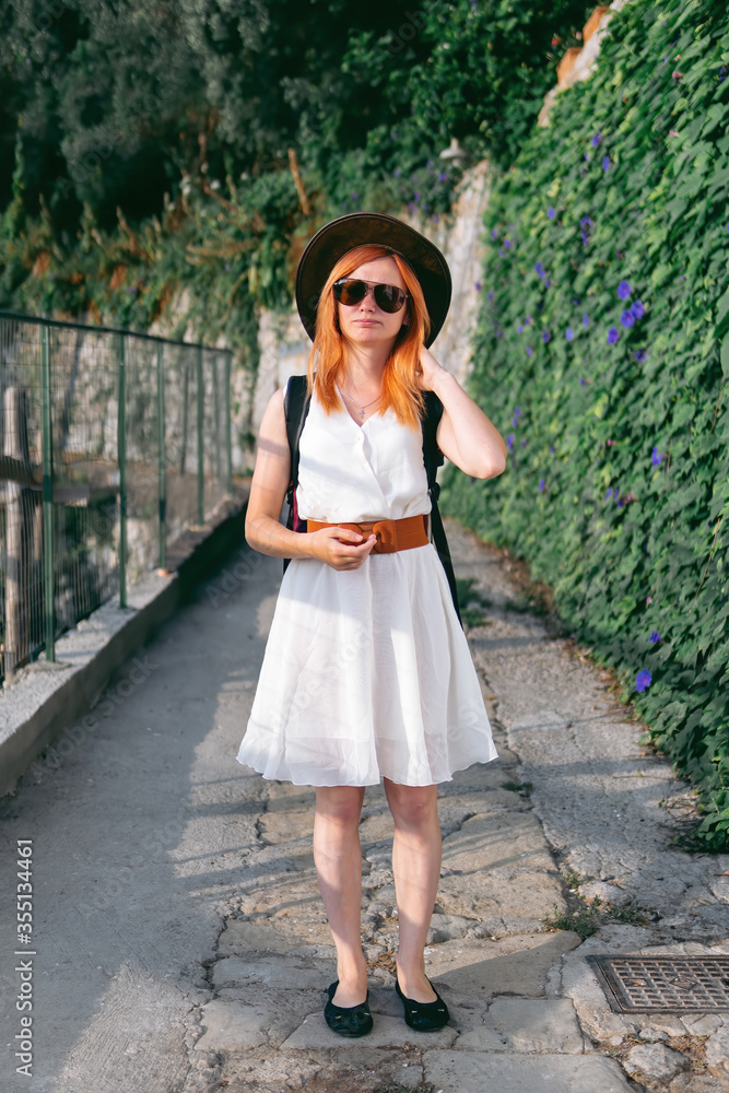 Summer day. A girl in a white dress and hat walks along the narrow streets of Italy. Backpack behind. Travel and fashion concept. Vacation and rest. Vertical photo. Front view