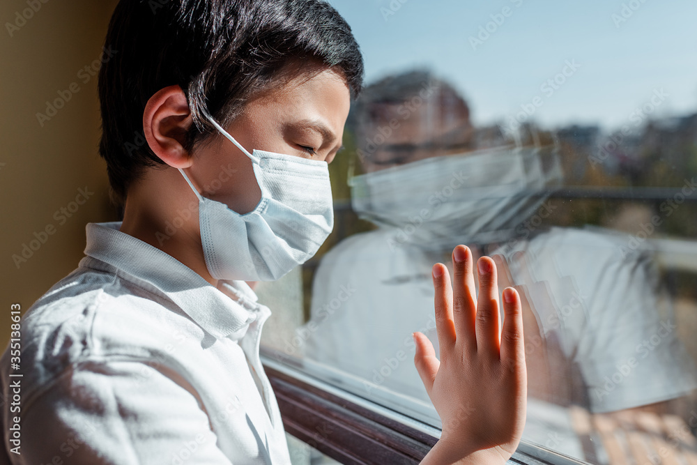 upset asian child in medical mask looking through window on quarantine upset asian child in medical mask looking through window on quarantine