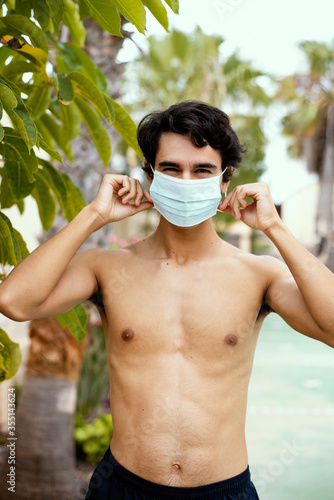 Handsome boy putting on surgery protective mask during the coronavirus pandemic in tropical environment with plants