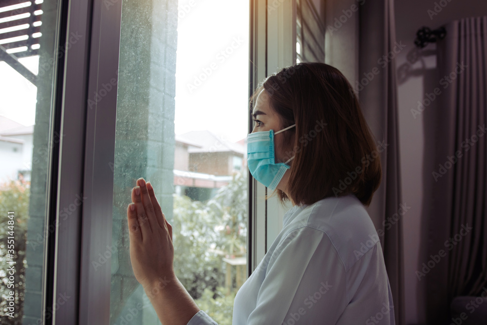 Asian woman looking through the window and wearing medical mask protection from the illness with stay isolated quarantine COVID-19 pandemic virus mask against coronavirus disease at home.